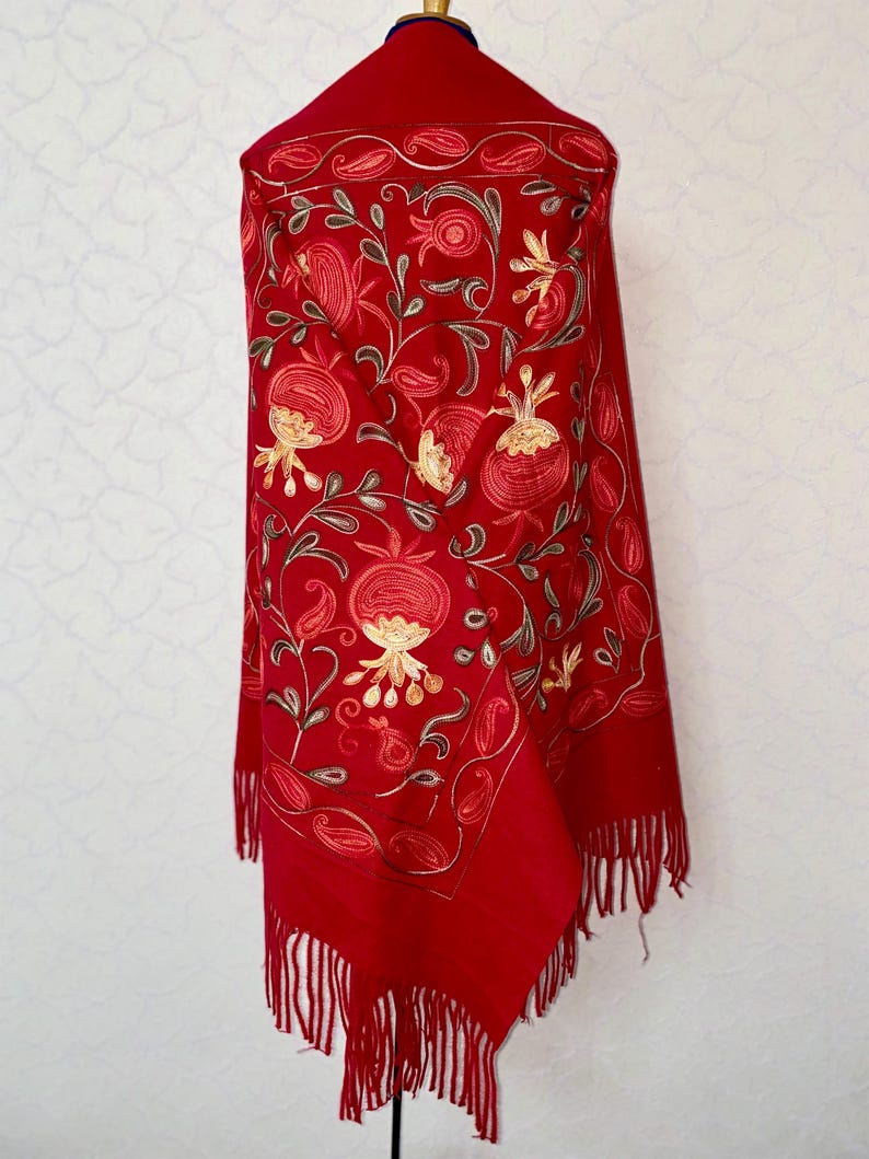 May include: A red shawl with embroidered floral designs in shades of gold, brown, and red. The shawl has fringe along the bottom edge.