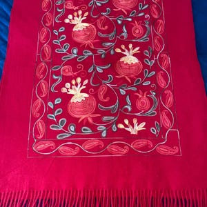 May include: A red wool shawl with embroidered pomegranate designs in shades of gold, green, and orange. The shawl has a fringe border.