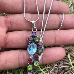 May include: A silver chain necklace with a pendant featuring a cluster of colorful gemstones. The pendant is shaped like a teardrop and features red, green, blue, and purple gemstones.