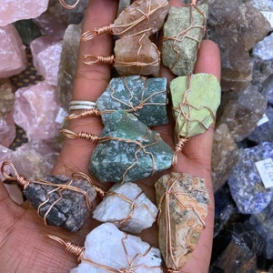 May include: A collection of 10 unique, raw, natural stone pendants wrapped in copper wire. The stones are various colors, including green, white, brown, and pink. Each pendant has a copper loop for hanging.