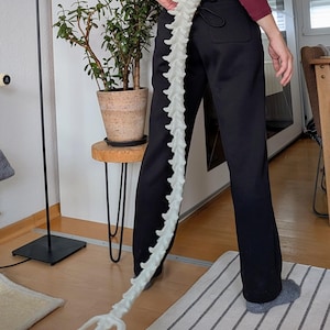 Op de afbeelding: Een witte, ruggengraatvormige kostuumaccessoire. De prop wordt vastgehouden door een persoon die een zwarte broek en een bordeauxrode top met lange mouwen draagt. De prop is gemaakt van een stijf materiaal en heeft een gestructureerd oppervlak. De staart is ongeveer 100 cm lang.