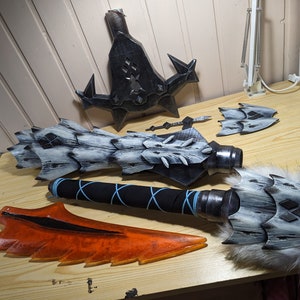 May include: A collection of fantasy weapon parts, including a large blade, a spiked shield, and a hammer head with fur trim. The pieces are painted in shades of gray, black, and orange, suggesting a dragon or monster theme.