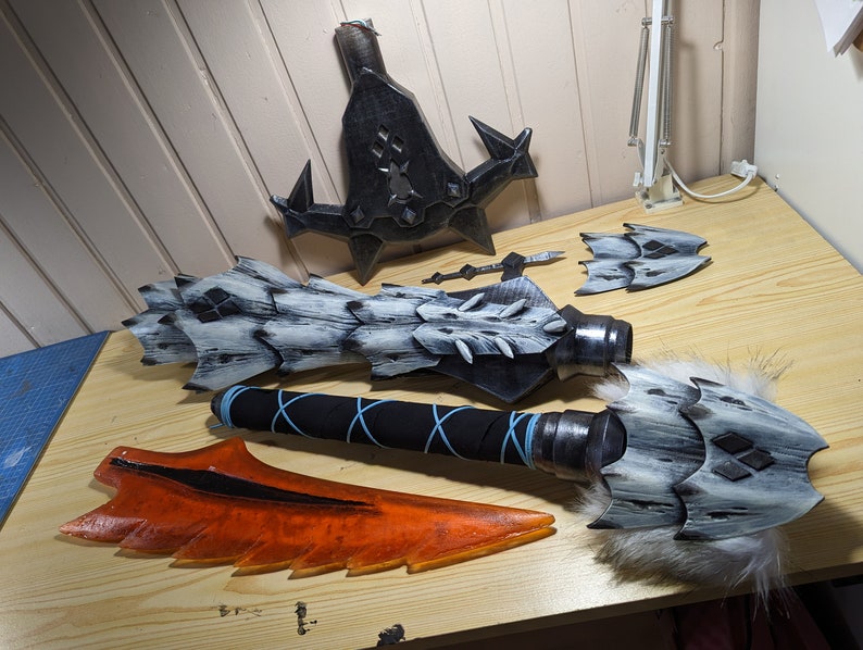 May include: A collection of fantasy weapon parts, including a large blade, a spiked shield, and a hammer head with fur trim. The pieces are painted in shades of gray, black, and orange, suggesting a dragon or monster theme.