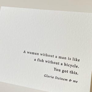 Peut inclure: Une carte rectangulaire blanche à la surface texturée présentant un texte imprimé en noir qui dit : A woman without a man is like a fish without a bicycle. You got this. Gloria Steinem & me. La carte est orientée horizontalement.