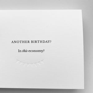 May include: A white greeting card featuring black printed text that reads ANOTHER BIRTHDAY? In this economy? Below the text is a subtle, embossed line drawing of a string of triangular bunting flags, creating a clean and minimalist design.
