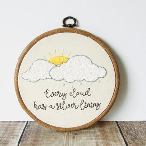 May include: Embroidered hoop art featuring a white cloud with a yellow sun peeking out and raindrops falling. The text "Every cloud has a silver lining" is embroidered below the cloud.