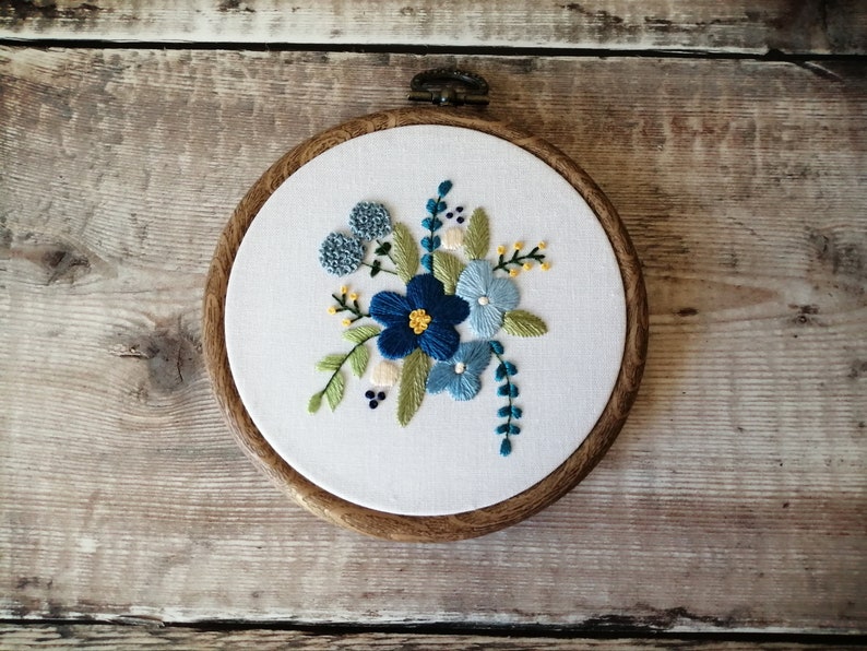 May include: Embroidered hoop art featuring a floral design with blue, green, and yellow flowers on a white background.