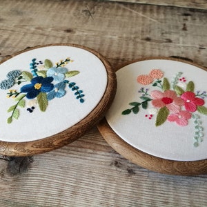 May include: Two embroidery hoops with floral designs. The hoop on the left has blue and green flowers. The hoop on the right has pink and green flowers.