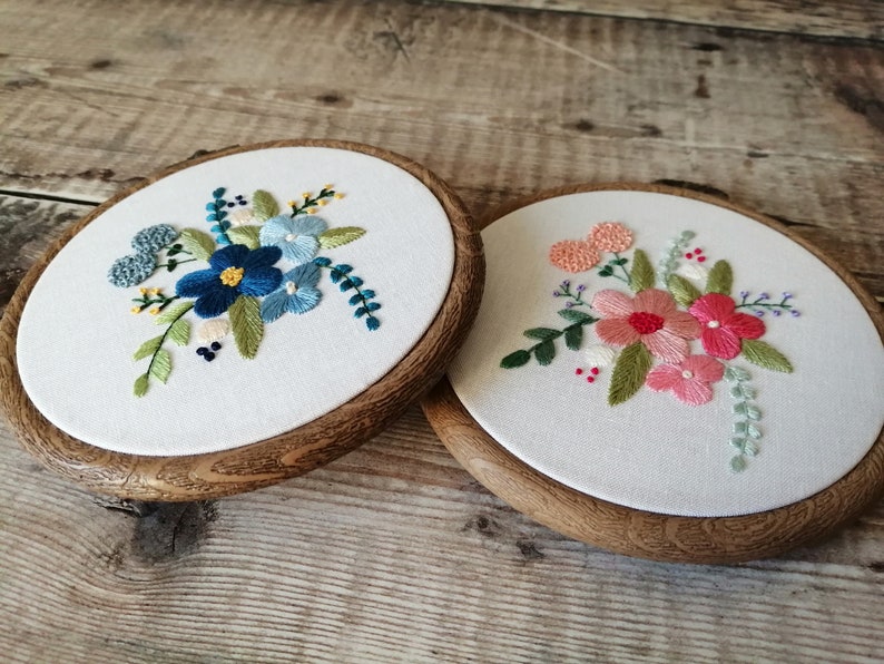 May include: Two embroidery hoops with floral designs. The hoop on the left has blue and green flowers. The hoop on the right has pink and green flowers.
