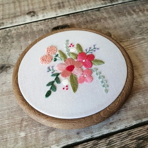 May include: A white embroidery hoop with a floral design. The flowers are pink, red, and white, and the leaves are green. The hoop is made of wood and has a natural finish.