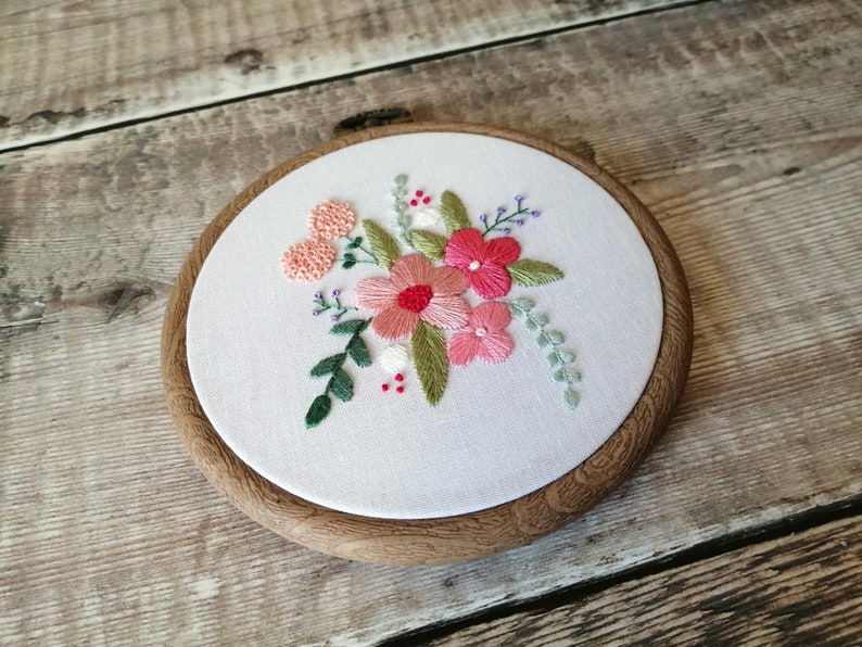 May include: A white embroidery hoop with a floral design. The flowers are pink, red, and white, and the leaves are green. The hoop is made of wood and has a natural finish.