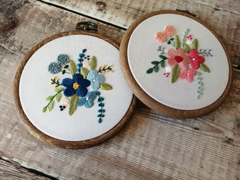 May include: Two embroidery hoops with floral designs. The hoop on the left features blue and green flowers, while the hoop on the right features pink and green flowers.