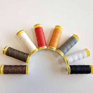 Könnte beinhalten: Acht Garnrollen in verschiedenen Farben, darunter Weiß, Grau, Braun, Schwarz, Orange und Rot. Die Rollen sind in einem Halbkreis auf einer weißen Oberfläche angeordnet.