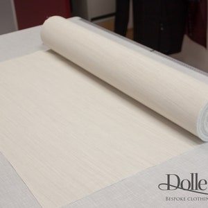 May include: A roll of white fabric with a subtle texture, unfurling on a gray surface. The fabric is labeled "Dolle Griet Bespoke Clothing and Costumes".