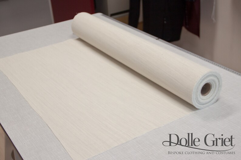 May include: A roll of white fabric with a subtle texture, unfurling on a gray surface. The fabric is labeled "Dolle Griet Bespoke Clothing and Costumes".