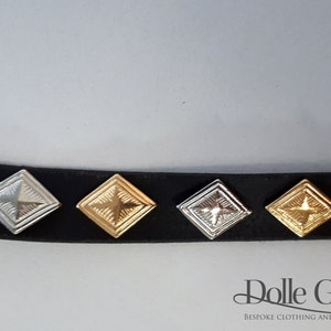 May include: Four square metal studs with a star design in different finishes: silver, gold, antique silver, and antique gold. The studs are attached to a black leather strap.