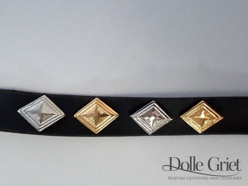 May include: Four square metal studs with a star design in different finishes: silver, gold, antique silver, and antique gold. The studs are attached to a black leather strap.
