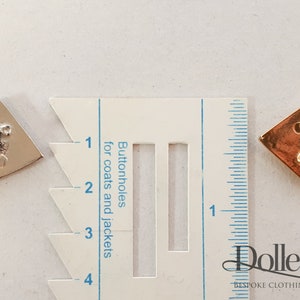 May include: Two diamond-shaped buttonhole cutters, one silver and one gold, are shown next to a ruler marked in inches. The ruler has a section labeled "Buttonholes for coats and jackets".