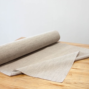 May include: A roll of natural beige linen fabric with a visible woven texture. The fabric is partially unrolled, revealing a flat section that extends across the surface. The material has a consistent light brown, earthy tone throughout the roll.