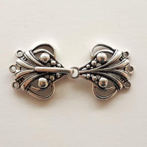 May include: Silver-coloured metal clasp with an ornate, symmetrical design. The clasp features curved lines, small spheres, and loops, connected by a central bar and ring. The decorative elements give it a vintage or antique aesthetic.