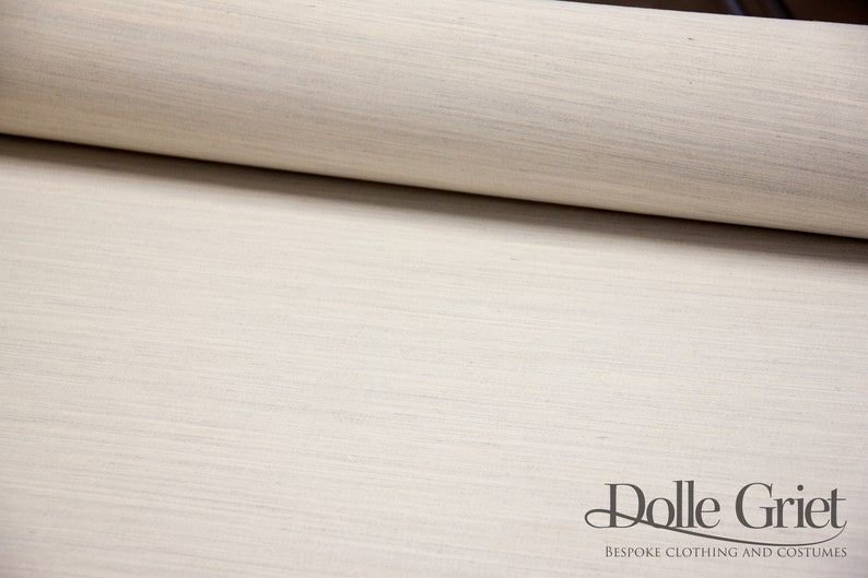 May include: Close-up of a roll of off-white fabric with a subtle woven texture. The fabric is smooth and has a slightly shiny finish. The text "Dolle Griet" is visible at the bottom of the image.