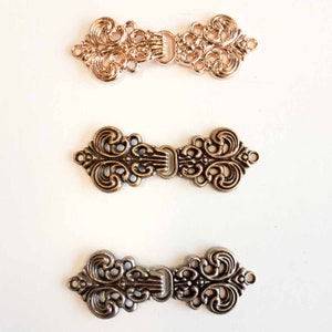 May include: Three ornate metal clasps in gold, bronze, and silver finishes. Each clasp features a decorative, intricate design with a central loop for attaching to fabric.
