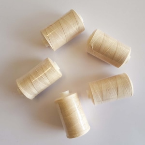 May include: Five spools of beige thread, arranged in a circle on a white surface.