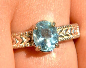 May include: A silver ring featuring an oval, light blue gemstone. The band has a detailed, patterned design. The ring is held by a person's finger, showcasing the jewelry's craftsmanship and color.