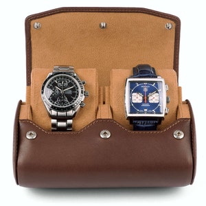 May include: A brown leather watch roll with two compartments for storing watches. The roll is open, revealing a silver chronograph watch with a black face and a silver square watch with a blue face.