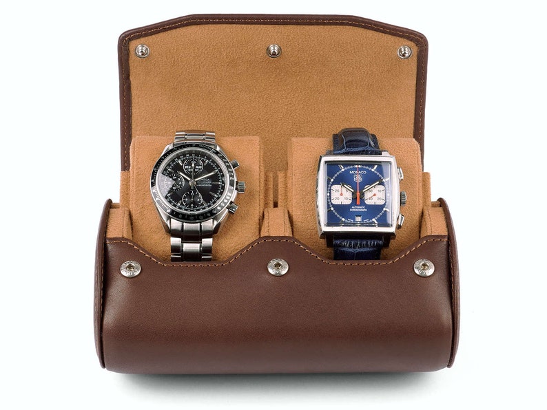May include: A brown leather watch roll with two compartments for storing watches. The roll is open, revealing a silver chronograph watch with a black face and a silver square watch with a blue face.
