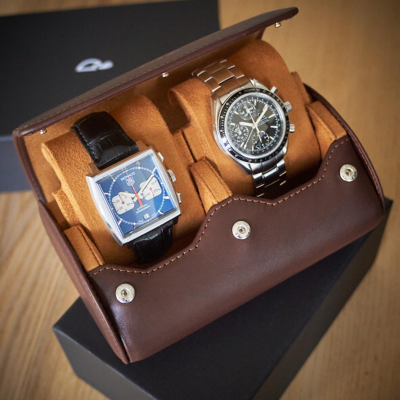 May include: A brown leather watch roll with two watches inside. The watch roll has a flap that closes with a snap. The watches are both chronograph watches with silver bands. One watch has a square face and the other has a round face.