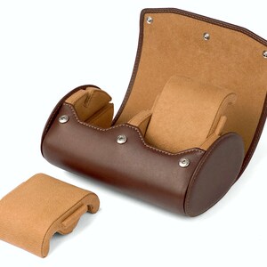 May include: A brown leather watch roll with a tan suede interior. The roll is open and the interior is visible. The roll has a separate compartment for a watch.