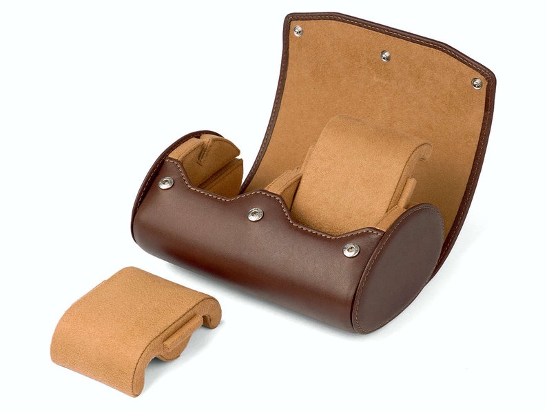 May include: A brown leather watch roll with a tan suede interior. The roll is open and the interior is visible. The roll has a separate compartment for a watch.