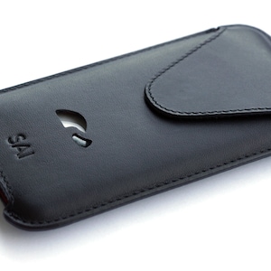 Peut inclure: Étui de téléphone en cuir noir avec une fermeture à rabat et une découpe pour l'appareil photo du téléphone. L'étui porte le mot "SAI" en relief sur le devant.