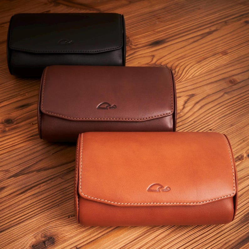 May include: Three leather watch roll cases in black, brown, and orange. Each case has a stitched design and a small logo on the front.