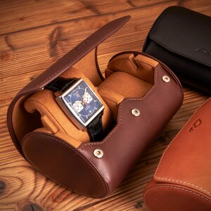 May include: A brown leather watch roll with a watch inside. The watch has a silver case and a blue face. The watch roll has a flap that can be closed to protect the watch.