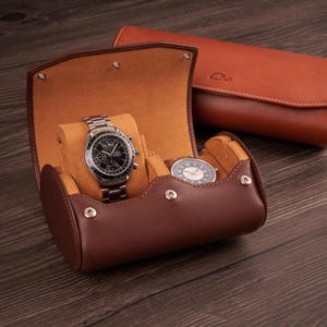 May include: A brown leather watch roll with two watch compartments. The roll is open and a silver watch with a black face is visible in one compartment. A second watch with a white face is visible in the other compartment.