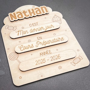 Op de afbeelding: Een houten plaat met de naam "Nathan" bovenaan. De plaat heeft drie tekstregels: "C'est Mon dernier jour", "EN Cours Préparatoire" en "ANNÉE 2025-2026". De plaat is versierd met gegraveerde snoepjes en sterren.