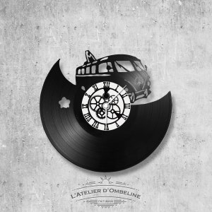 May include: Black vinyl record clock with a silhouette of a vintage van and clock gears. The clock face is white with black Roman numerals. The clock is mounted on a gray concrete wall. The text "L'ATELIER D'OMBELINE" and "FAIT-MAIN" is visible below the clock.