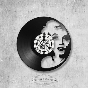 May include: Black vinyl record clock with a silhouette of a face and a white clock face with Roman numerals and gears. The clock has the text "L'ATELIER D'OMBELINE" and "FAIT-MAIN" on the bottom.