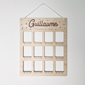 Peut inclure: Un cadre photo en bois avec le texte "Guillaume Ma premiere annee" et 12 emplacements photo vides pour chaque mois de l'année. Le cadre est décoré d'étoiles.