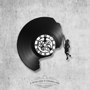May include: A black vinyl record clock with a white clock face and gears. The record is cut out to show a silhouette of a person climbing a rock face. The clock face reads "L'ATELIER D'OMBELINE - FAIT-MAIN".