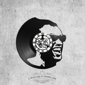 May include: Black vinyl record clock featuring a silhouette of a person wearing sunglasses. The clock face is a white circle with black Roman numerals and gears. The text "L'ATELIER D'OMBELINE" and "FAIT-MAIN" is printed below the clock.