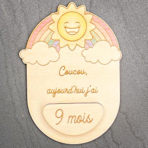 May include: Wooden baby milestone plaque with a smiling sun, rainbow, and clouds design. The plaque has the text "Coucou, aujourd'hui j'ai 9 mois" engraved on it. The plaque is light brown in color.