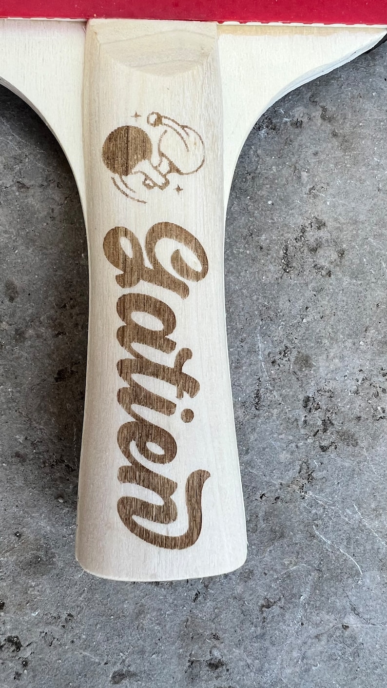 May include: A wooden table tennis paddle with a red surface and a white handle. The handle has a graphic of a person playing table tennis and the word "Gatlin" engraved on it.