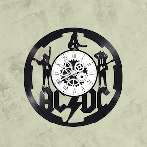 Könnte beinhalten: Eine schwarze Schallplattenuhr mit dem AC/DC-Logo und Silhouetten der Bandmitglieder. Das Zifferblatt der Uhr ist weiß mit schwarzen römischen Ziffern als Stundenmarkierungen und schwarzen, zahnradförmigen Zeigern.