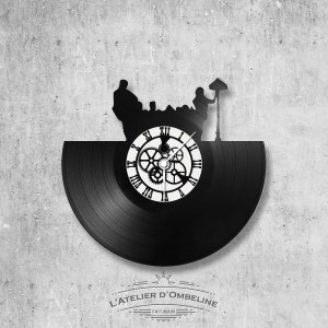 May include: Black vinyl record clock with a silhouette of three people sitting at a table with a street lamp. The clock face features Roman numerals and gears. The text "L'ATELIER D'OMBELINE - FAIT-MAIN" is printed below the clock.