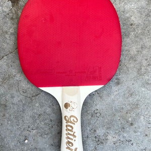 May include: A red and white table tennis paddle with a wooden handle. The handle has the word "Gatien" engraved on it.
