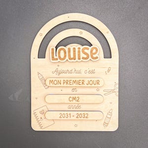 Op de afbeelding: Een houten bord met de naam "Louise", met daaronder secties voor "MON PREMIER JOUR" en het schooljaar 2031-2032. Versierd met potlood- en schaar illustraties en een regenboogboog ontwerp.