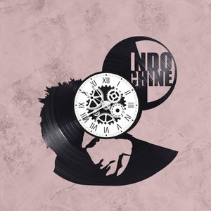 May include: A black vinyl record clock with a silhouette of a person's head and the text "INDO CHINE" on the face. The clock has a white face with black Roman numeral hour markers and black gear-shaped hands.